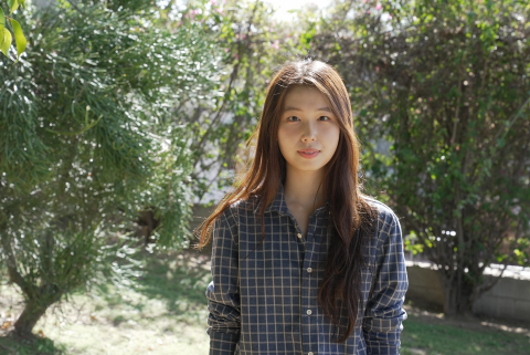 Woman with long hair, smiling, standing in front of green trees.