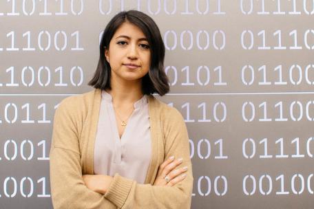upper body photograph of a woman with her arms crossed in front of a wall showing binary code
