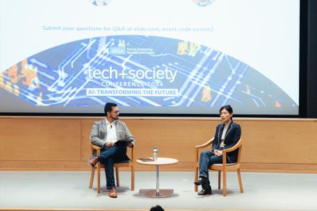 Two people sitting on stage having an event discussion in front of an auditorium screen 
